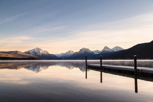 Serene lake at sunrise, reflecting snow-capped mountains and a wooden dock in calm waters.