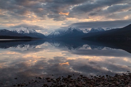 A serene lake reflects snow-capped mountains and a cloudy sky at sunset, with pebbles in the foreground.