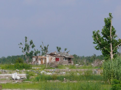 A damaged house surrounded by debris and fallen trees, set against a clear blue sky.