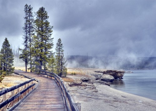A wooden walkway leads through misty trees along a serene lakeshore, with rocky cliffs in the background.