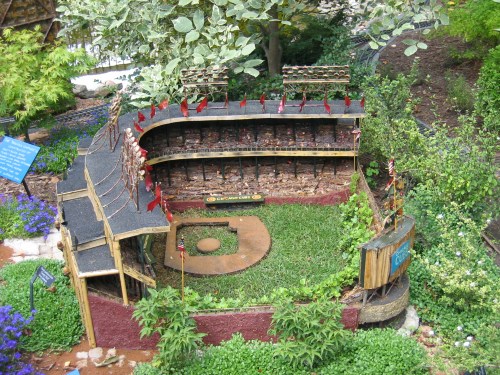 A miniature baseball stadium surrounded by greenery and flowers, featuring seating and a small field.