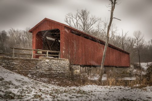 A red covered bridge stands in a snowy landscape, surrounded by bare trees and a stone foundation.