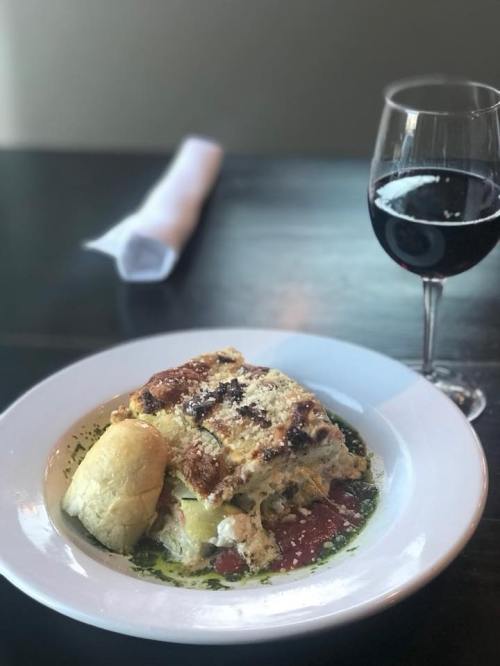 A plate of lasagna with a side roll, garnished with sauce, next to a glass of red wine on a dark table.