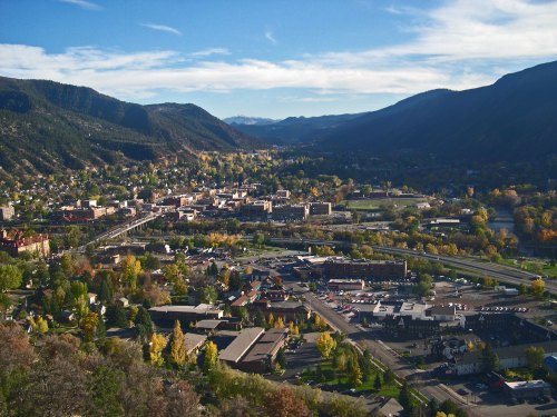 Aerial view of a town surrounded by mountains, featuring colorful autumn trees and a clear blue sky.