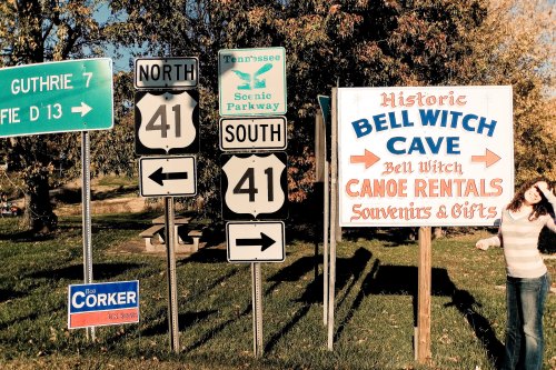 A woman stands next to road signs for Bell Witch Cave and Canoe Rentals, with directional signs for Guthrie and Tennessee.