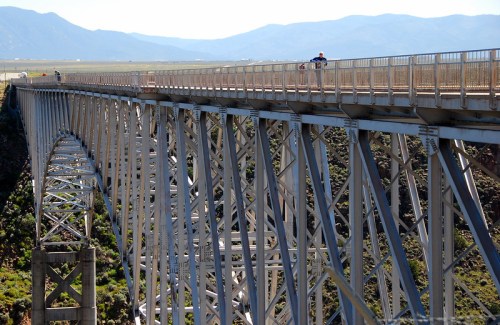 A long, metal bridge spans a canyon, with people walking along its railing against a backdrop of mountains.