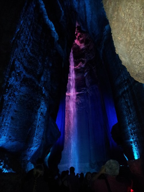 A stunning cave interior with colorful lighting and a waterfall cascading down the rock walls, surrounded by visitors.