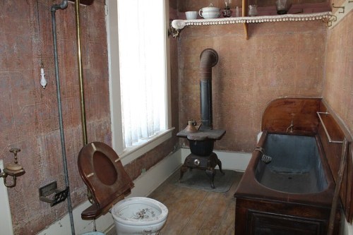 A vintage bathroom featuring a wooden bathtub, a stove, and an old-fashioned toilet against patterned wallpaper.