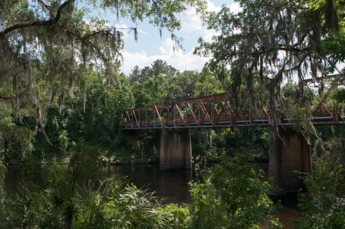 A rusted metal bridge spans a calm river, surrounded by lush greenery and hanging moss under a bright blue sky.
