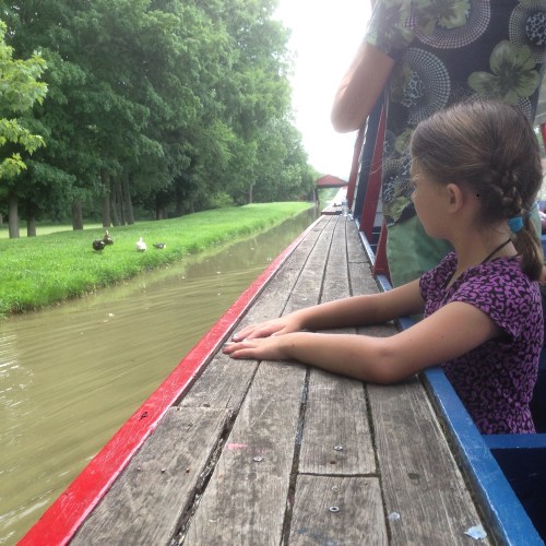 A girl sits on a boat, watching ducks along a calm, tree-lined waterway on a sunny day.