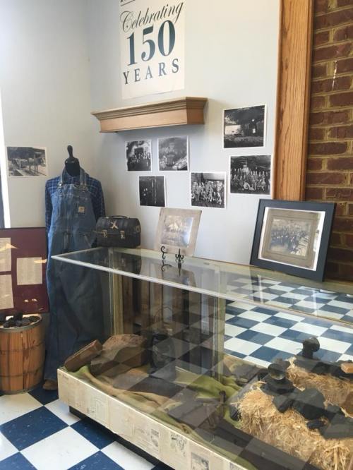 Display case with historical artifacts, a mannequin in overalls, and black-and-white photos on the wall. Celebrating 150 years.
