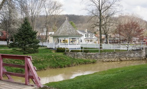 A quaint gazebo by a creek, surrounded by trees and buildings, in a peaceful rural setting.
