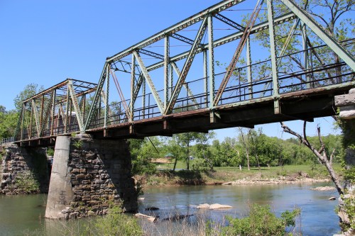A weathered green metal bridge spans a calm river, surrounded by lush greenery and clear blue skies.