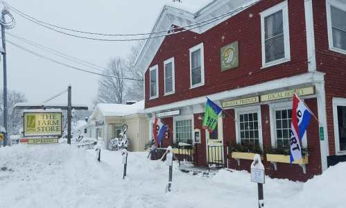 A red farm store building covered in snow, with colorful flags and signs for local produce and honey.