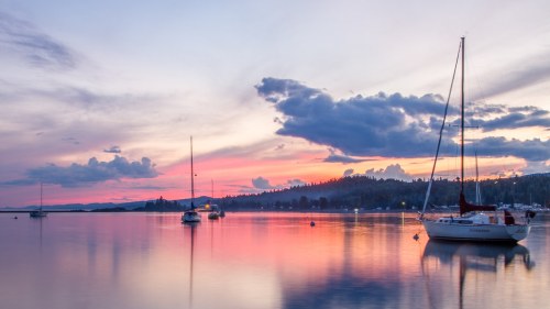 A serene sunset over a calm lake, with sailboats anchored and colorful clouds reflecting on the water.