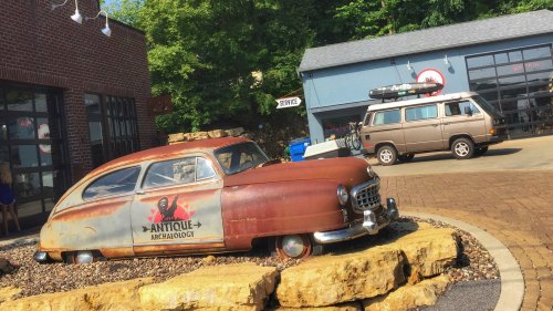 Rusty vintage car with "Antique Archaeology" sign, parked on rocky ground near a building and another vehicle in the background.