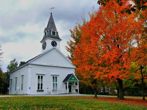 A white church with a clock tower beside a vibrant orange tree in autumn, under a cloudy sky.