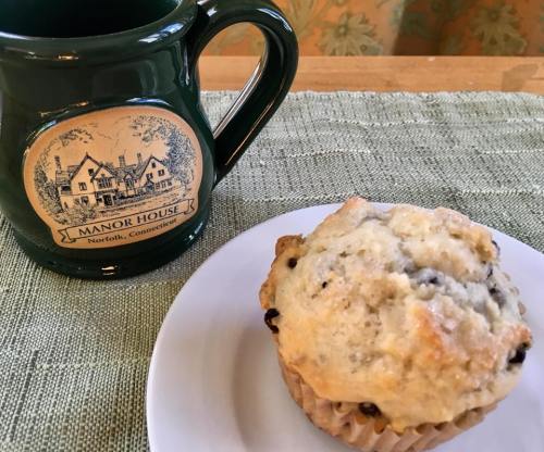 A green mug with "Manor House" design next to a plate with a muffin on a textured green tablecloth.