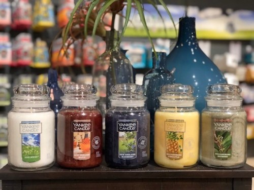 Five Yankee Candle jars in various colors, displayed on a shelf with decorative vases in the background.