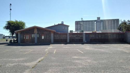 A vacant building with a wooden facade and a large grain elevator in the background, set in a gravel parking lot.