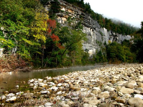 A serene riverbank with smooth stones, surrounded by colorful trees and towering cliffs, shrouded in morning mist.