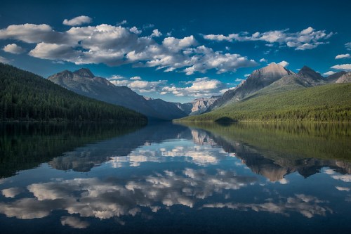 A serene lake reflects mountains and clouds under a blue sky, surrounded by lush green forests.
