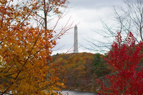 A tall monument rises above colorful autumn trees by a calm lake under a cloudy sky.
