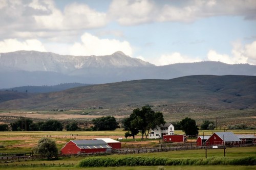 A scenic view of red barns and a farmhouse against rolling hills and mountains under a cloudy sky.