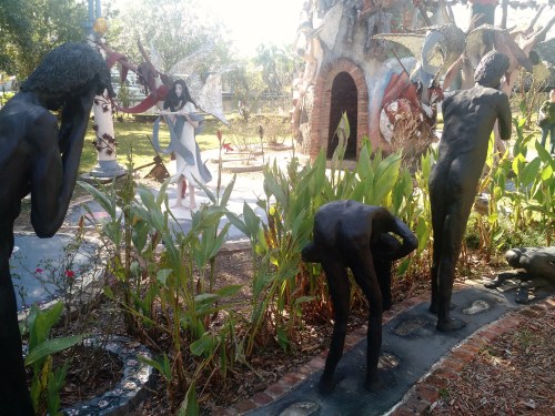 A garden with various sculptures, including figures in different poses surrounded by greenery and artistic structures.