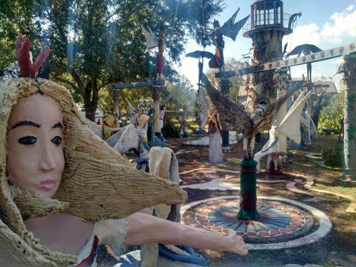 A whimsical outdoor art installation featuring colorful sculptures of angels and birds among trees and a lighthouse.