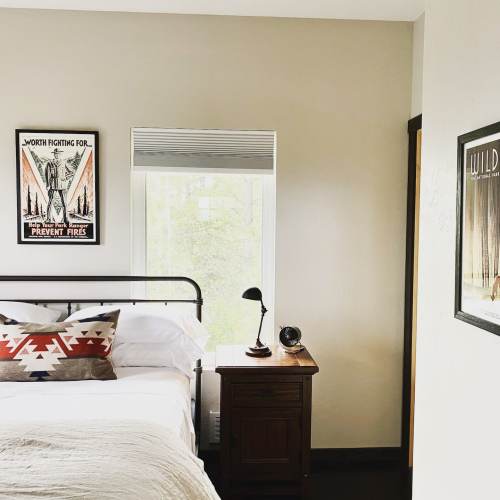 A cozy bedroom featuring a bed, bedside table with a lamp, and framed artwork on the walls. Natural light from a window.