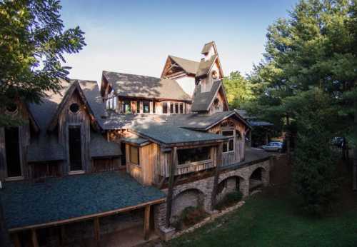 A large, rustic-style house with multiple gables, surrounded by trees and a stone foundation.
