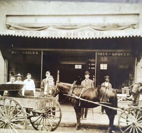 Historic photo of a grocery store with a horse-drawn cart and several men standing outside the entrance.