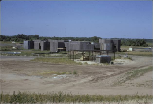 A large, modern building complex under construction in a rural area, surrounded by open land and blue sky.
