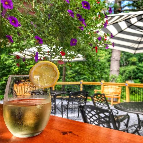 A glass of white wine with a lemon slice sits on a wooden table, surrounded by vibrant flowers and outdoor seating.