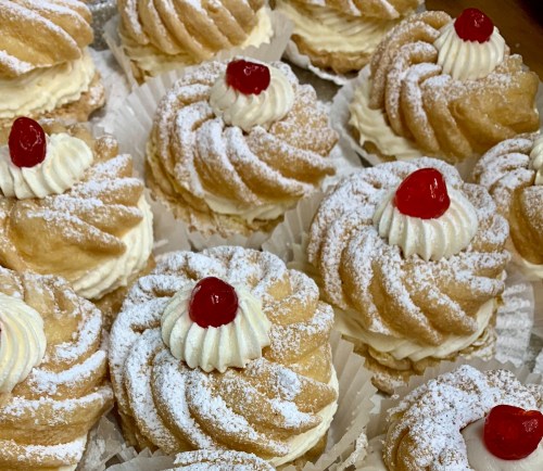 A close-up of cream-filled pastries topped with whipped cream and cherries, dusted with powdered sugar.