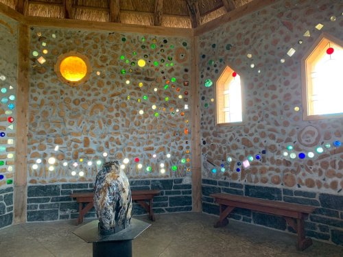 Interior of a stone building with colorful glass pieces embedded in the walls and a central sculpture on a pedestal.