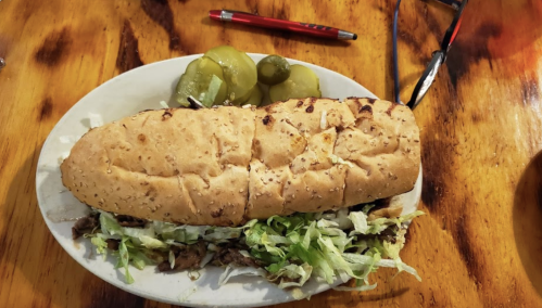 A large sandwich with sesame seed bread, filled with lettuce and meat, served with pickles on the side.