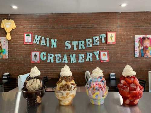 Four colorful ice cream sundaes with whipped cream sit on a counter, with a "Main Street Creamery" sign in the background.