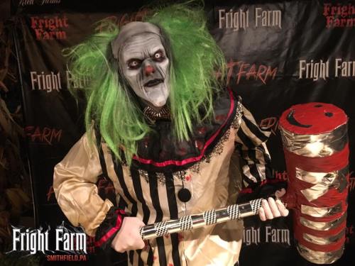 A creepy clown with green hair and a striped outfit holds a large mallet, standing in front of a Fright Farm backdrop.