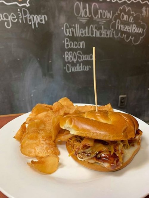 A grilled chicken sandwich on a pretzel bun with BBQ sauce and cheese, served with a side of potato chips.