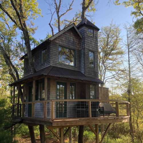 A charming treehouse with a wooden deck, surrounded by trees and greenery, featuring large windows and a rustic design.