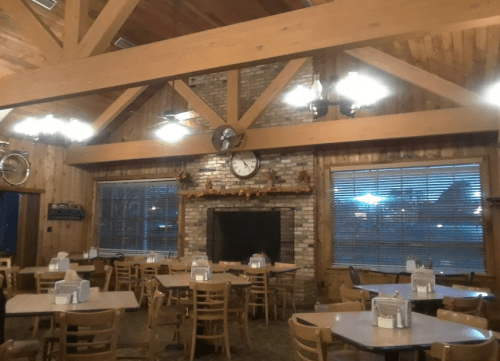 Cozy wooden interior of a restaurant with tables, a stone fireplace, and large windows, decorated with autumn leaves.