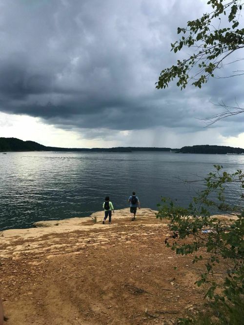 Two people walk along a rocky shore under a cloudy sky, with a calm lake and distant hills in the background.