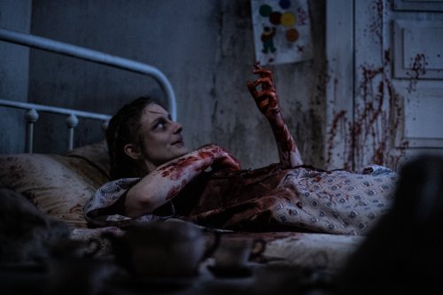 A woman lies in bed, covered in blood, with a distressed expression in a dimly lit, messy room.