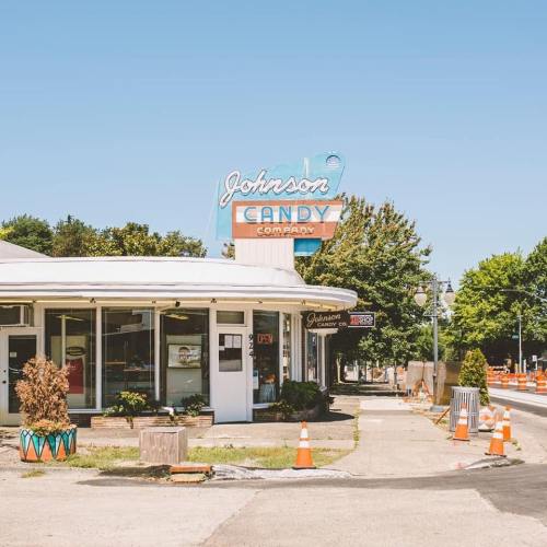 A vintage candy shop with a neon sign, surrounded by trees and road construction cones on a sunny day.