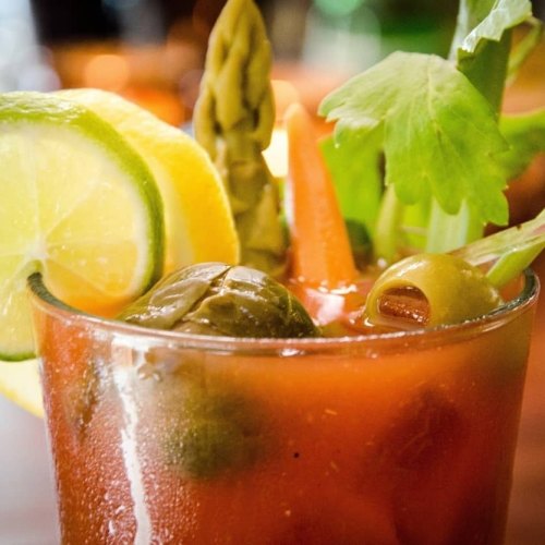 A close-up of a colorful cocktail garnished with lemon, lime, asparagus, celery, olives, and a carrot.