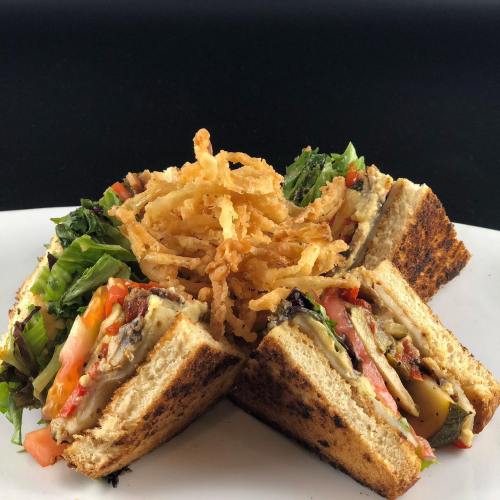 A stacked sandwich with layers of vegetables, served with crispy onion rings on top, on a white plate against a black background.