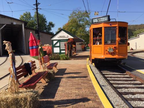 An orange tram at a station with autumn decorations, benches, and a nearby building in a sunny setting.