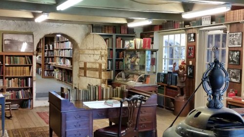 A cozy library interior with wooden shelves, a desk, and a display case, featuring books and framed photos on the walls.